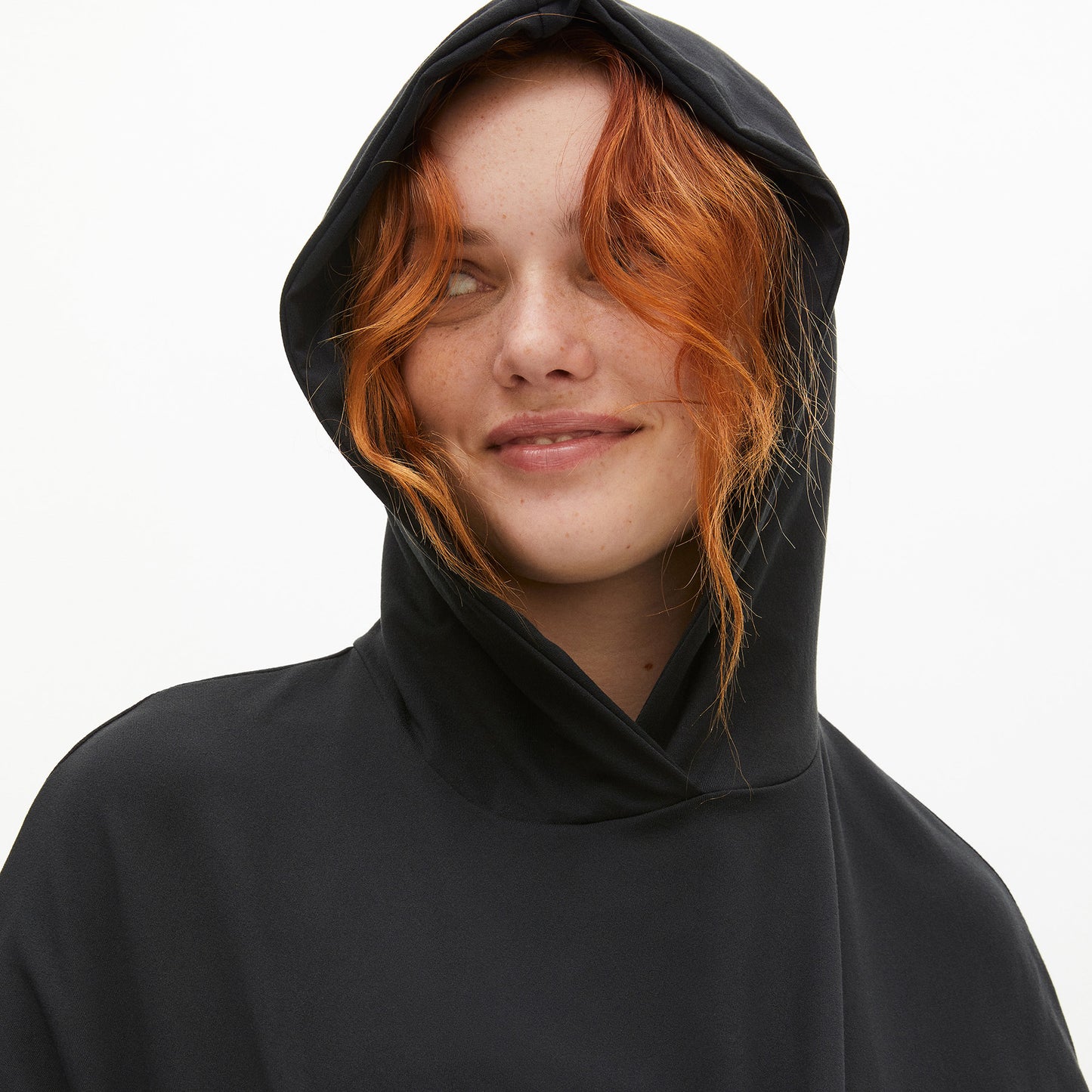 A red-haired person smiles warmly in the sleek Soft Jersey Hoodie by Rohnisch, made from recycled polyester, against a plain white backdrop.