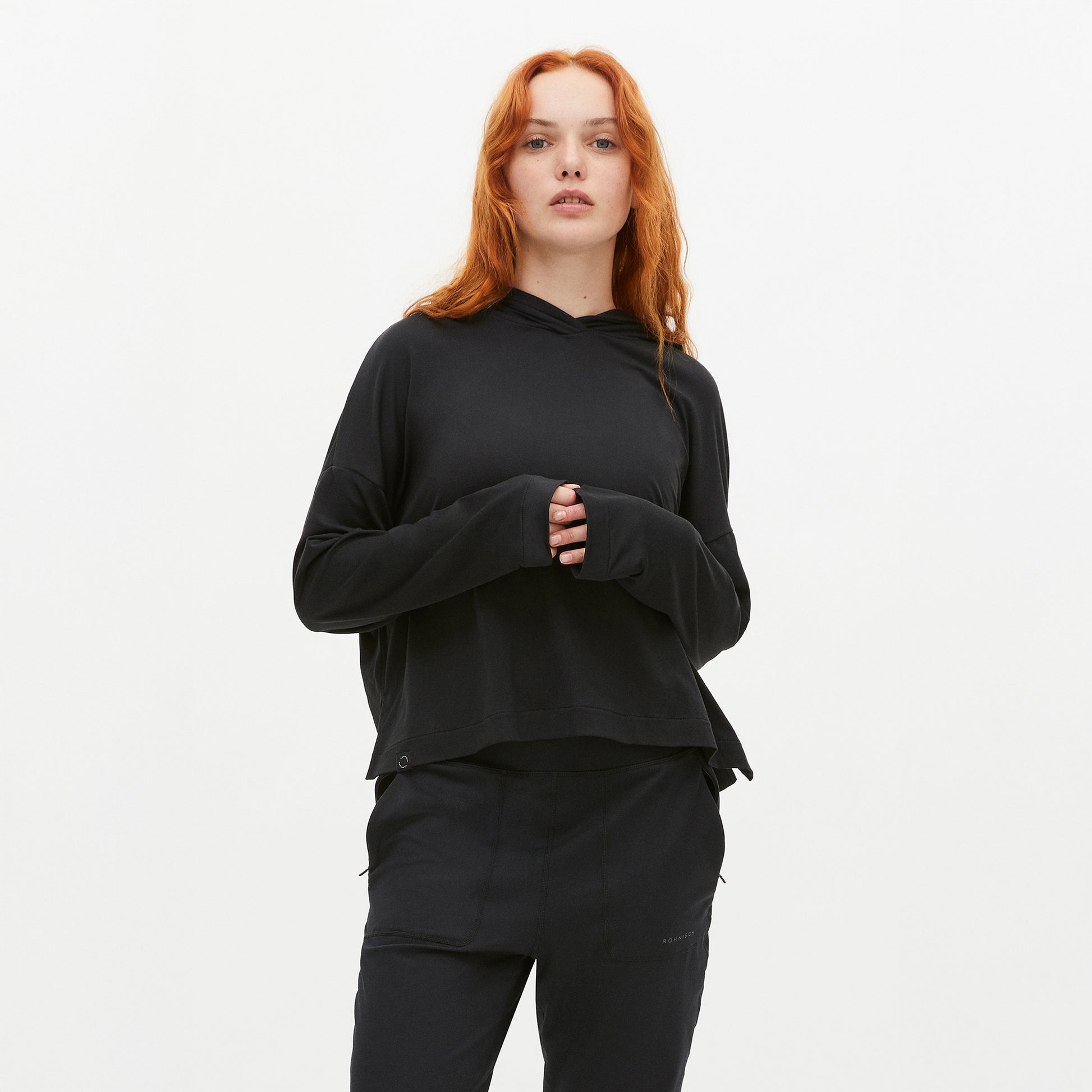 A person with long red hair wears a Rohnisch Soft Jersey Hoodie in black, made from recycled polyester, paired with black pants against a plain white background.
