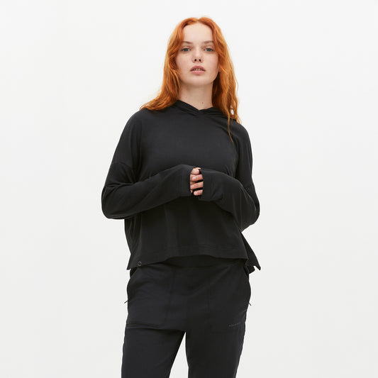 A person with long red hair wears a Rohnisch Soft Jersey Hoodie in black, made from recycled polyester, paired with black pants against a plain white background.
