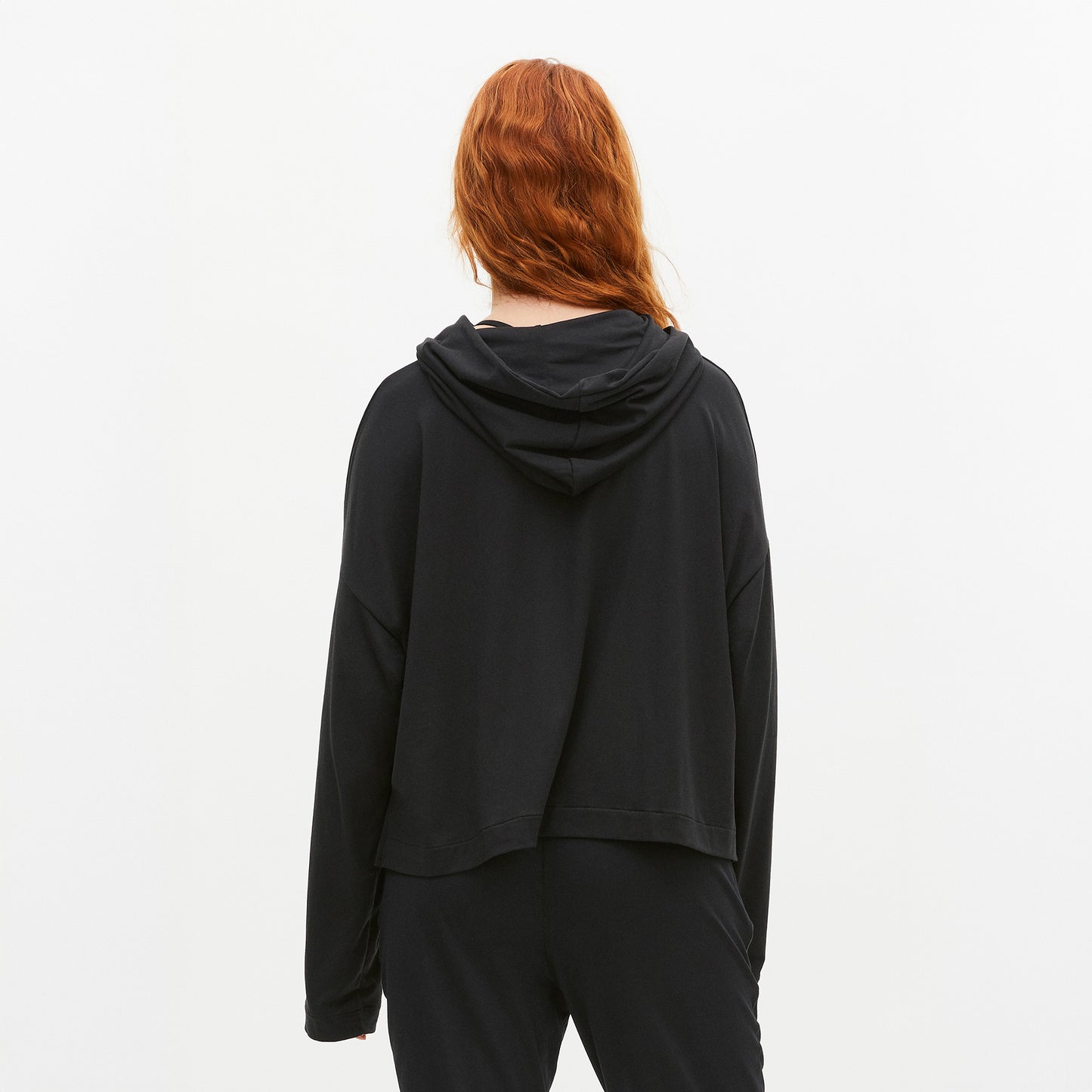 A person with long red hair is shown from the back, wearing a black Soft Jersey Hoodie and pants made from recycled polyester by Rohnisch against a plain white background.