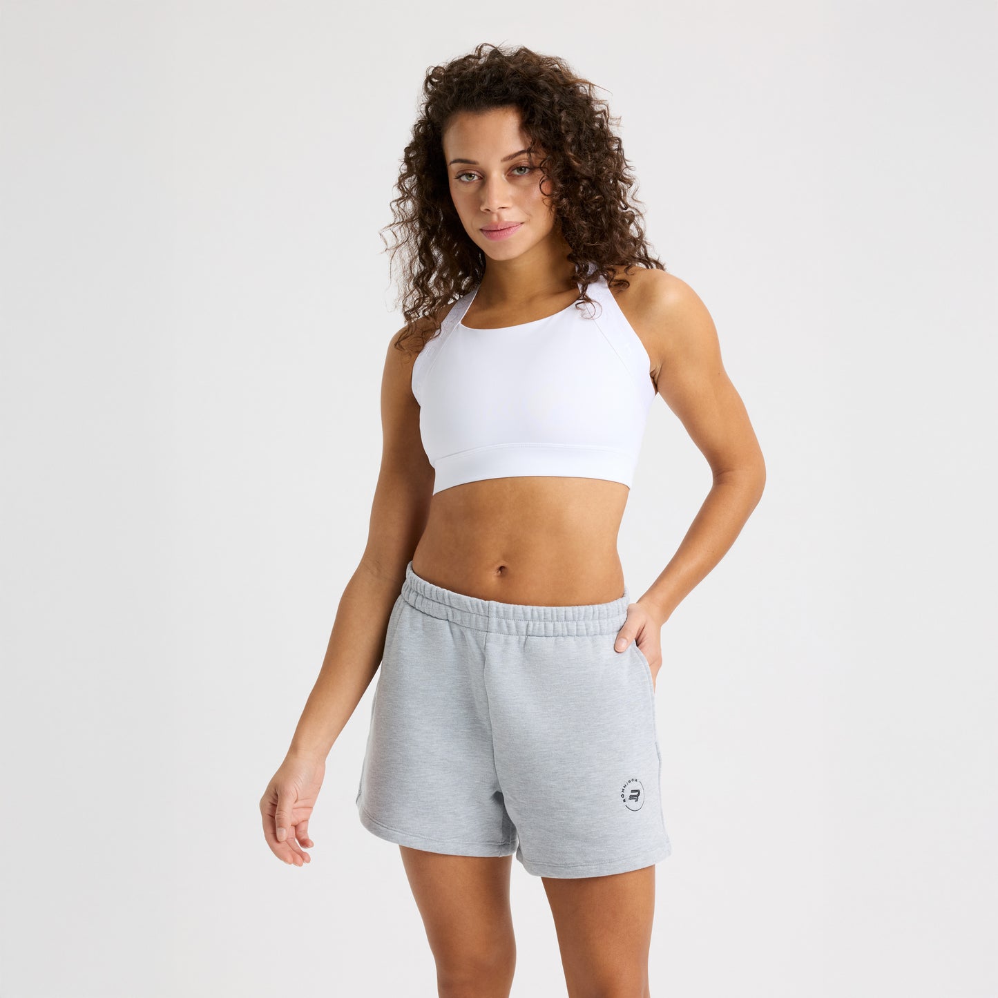 A person wears a Rohnisch white sports bra and Soft Sweatshort in gray, standing against a plain background, looking at the camera with one hand by their side. The soft terry fabric adds comfort, while a subtle logo print completes the look.
