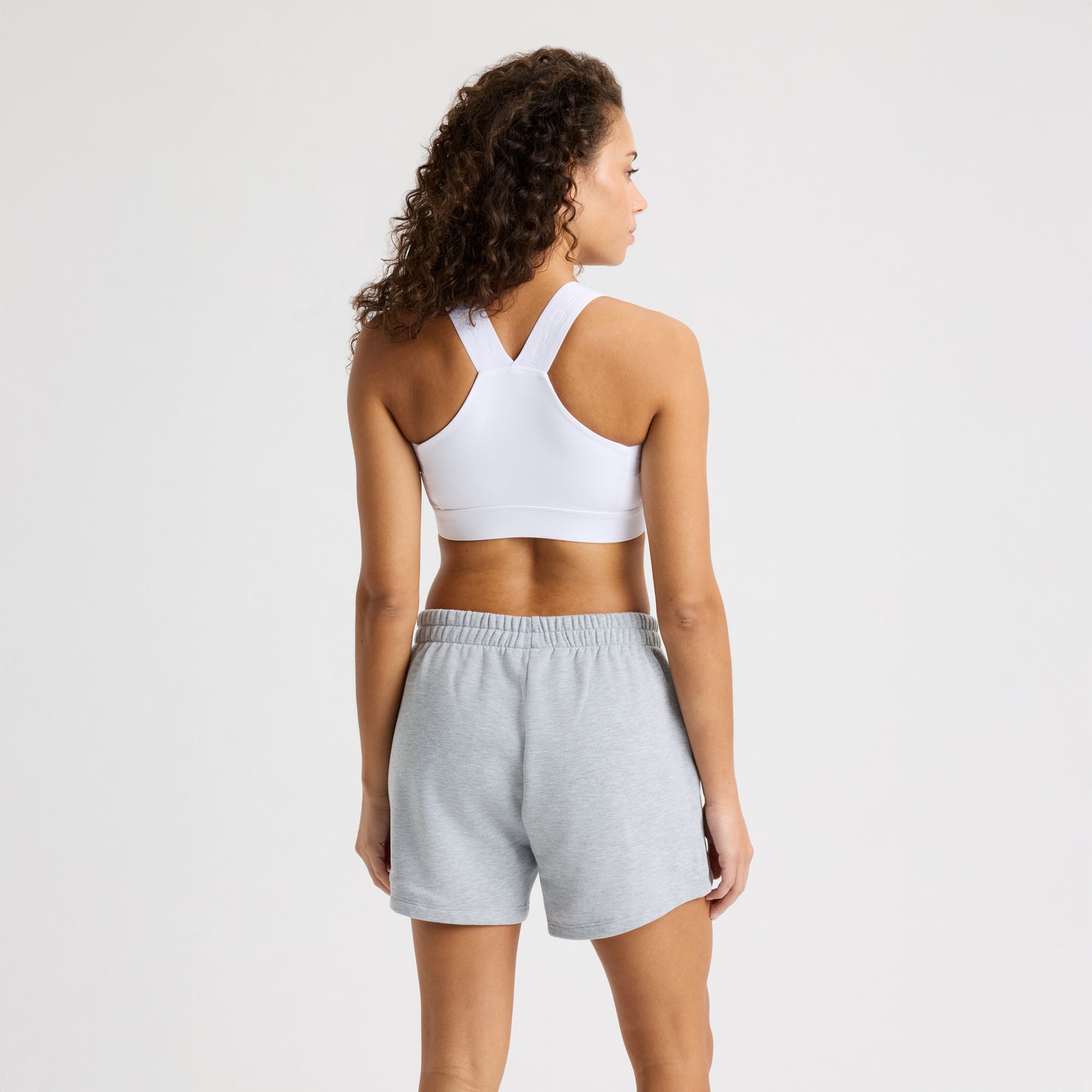 A person with curly hair in a white sports bra and gray Soft Sweatshort from Rohnisch faces away from the camera against a plain background.