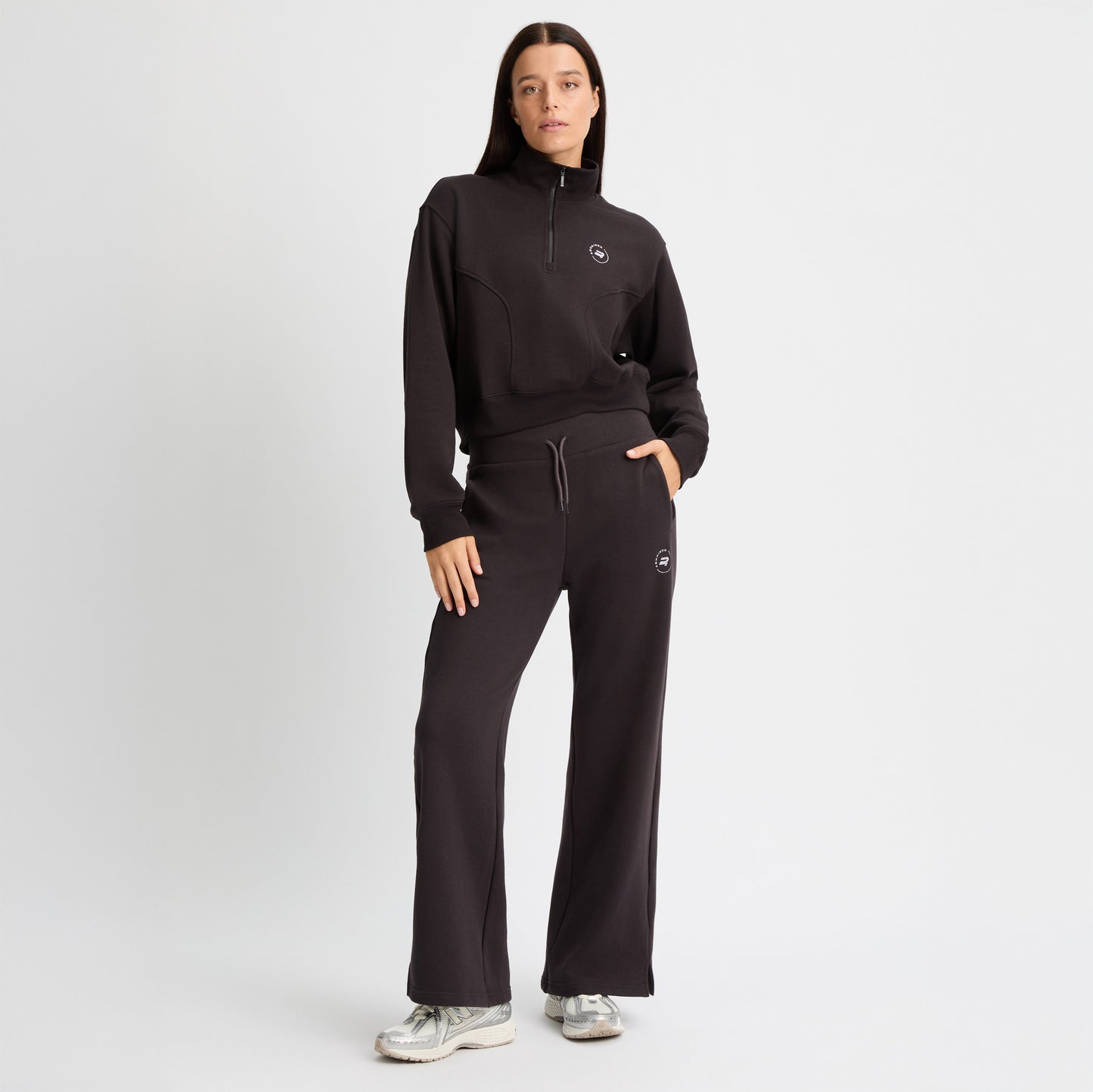 A woman in a black zip-up sweatshirt made from recycled fabric and Rohnisch's Soft Sweat Wide Pants stands with one hand in her pocket against a plain white background.