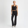 The individual in black athletic attire with gray sneakers poses against a white backdrop, highlighting Rohnisch's Ribbed Flair High Waist Tights made from recycled polyester, boasting a flared silhouette to complement their sleeveless top and pants look.