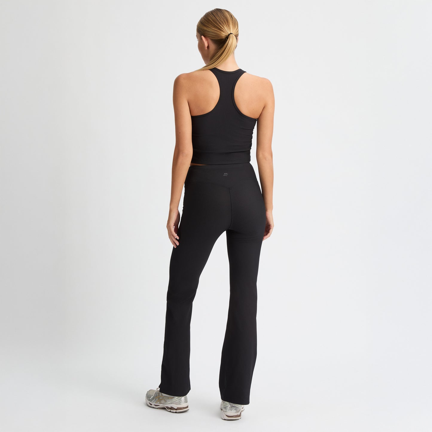 A woman with a blonde ponytail stands against a white background wearing a black athletic tank top and Rohnisch's Ribbed Flair High Waist Tights made from recycled polyester, facing away from the camera.