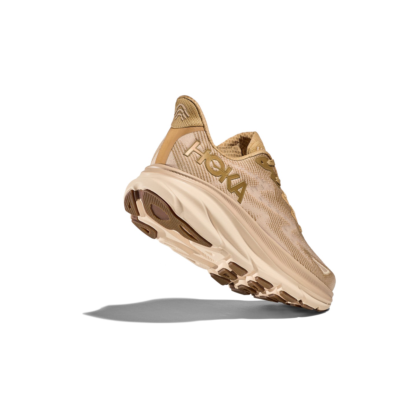 A Hoka M Clifton 9 running shoe in Wheat/Shifting Sand is shown in side profile against a white background, highlighting its cushioned sole and tread, with a shadow cast below.