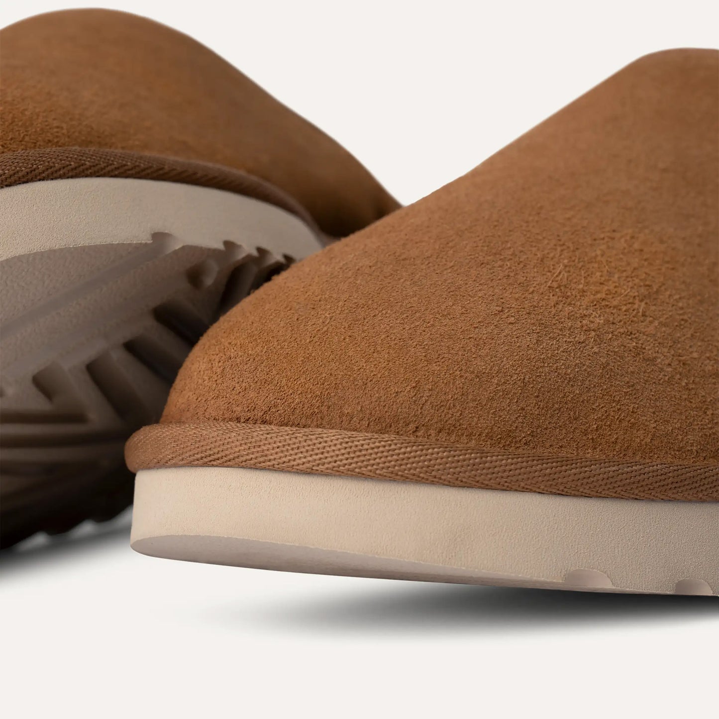 Close-up of two tan Ugg M Classic Slip-On slippers showcasing luxurious UGGplush™ lining and thick white rubber soles on a white background.