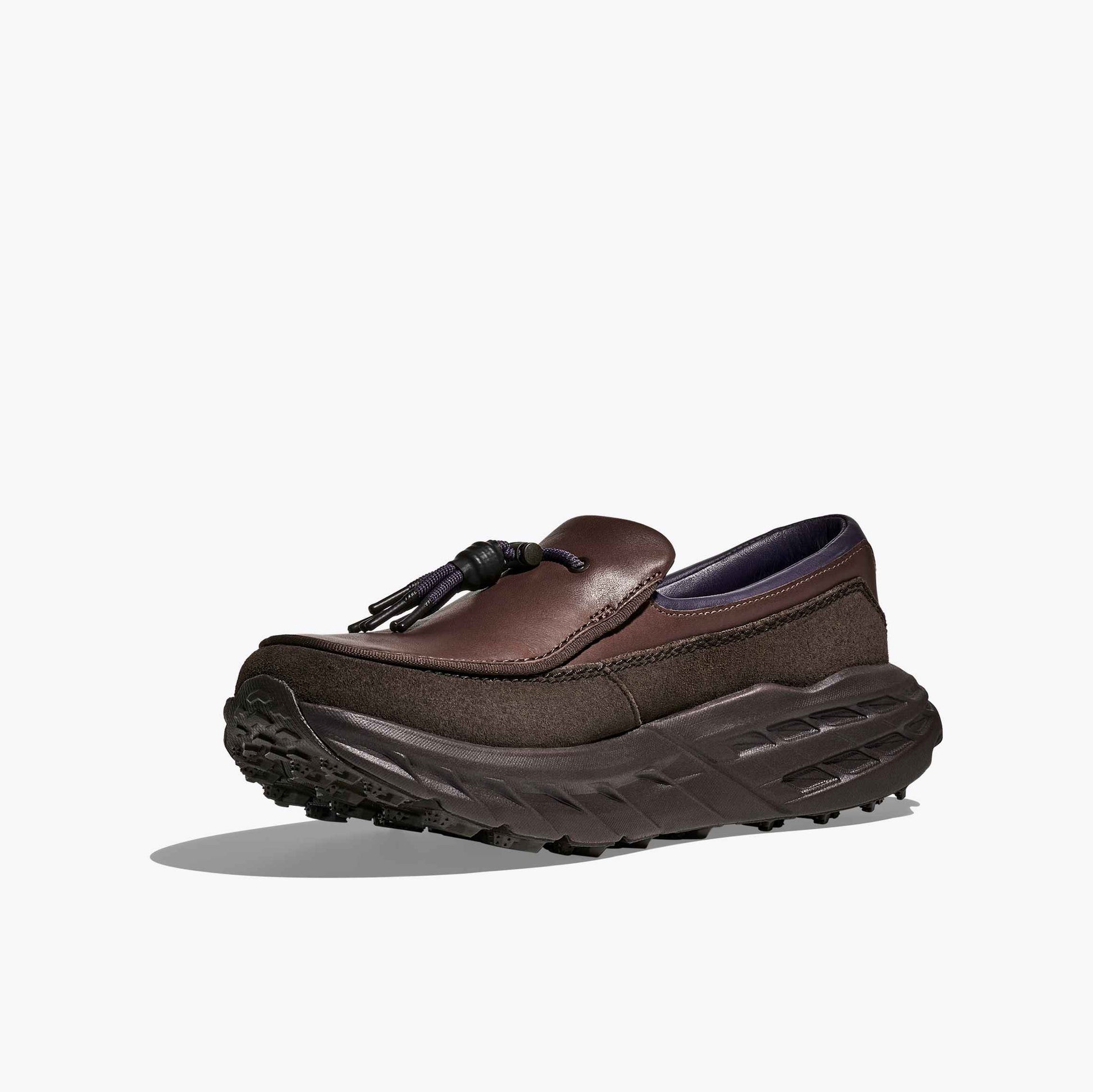 The Hoka U Speed Loafer is a brown luxury loafer featuring a thick, rugged black sole and tassel detail on the upper, shown against a plain white background.