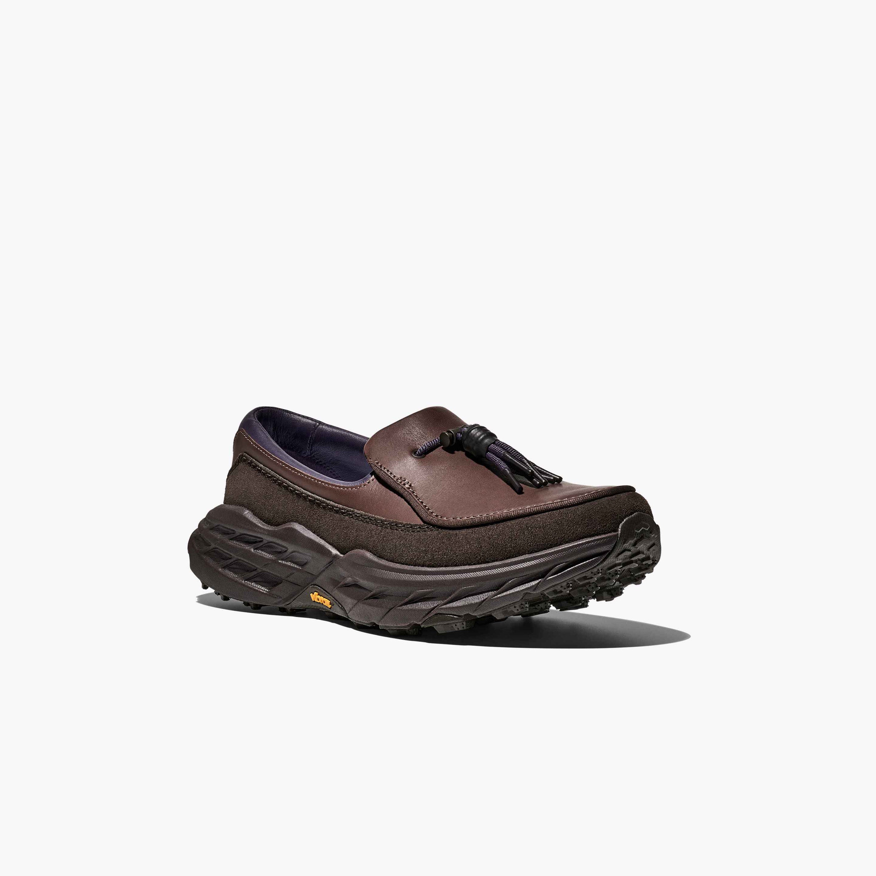 The Hoka U Speed Loafer is a dark brown leather and suede loafer with tassels, set on a thick textured black sole, shown against a plain white background.