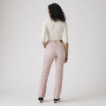 A person with curly hair, dressed in a white long-sleeve top, Levis 501 Original Scraped Pale Mauve jeans, and black sneakers, stands facing away against a plain background.
