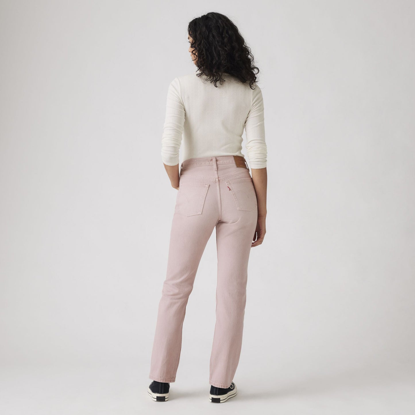 A person with curly hair, dressed in a white long-sleeve top, Levis 501 Original Scraped Pale Mauve jeans, and black sneakers, stands facing away against a plain background.