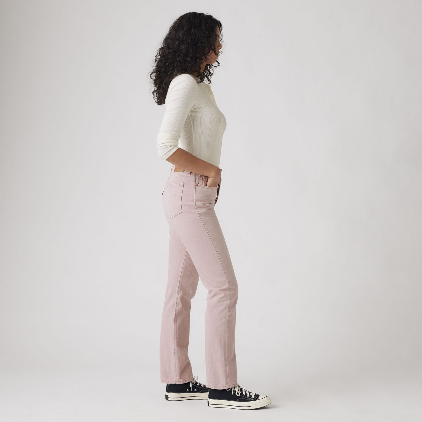 A person stands in profile against a plain background, wearing a white long-sleeve top, Levis 501 Original jeans in Scraped Pale Mauve, and black sneakers.