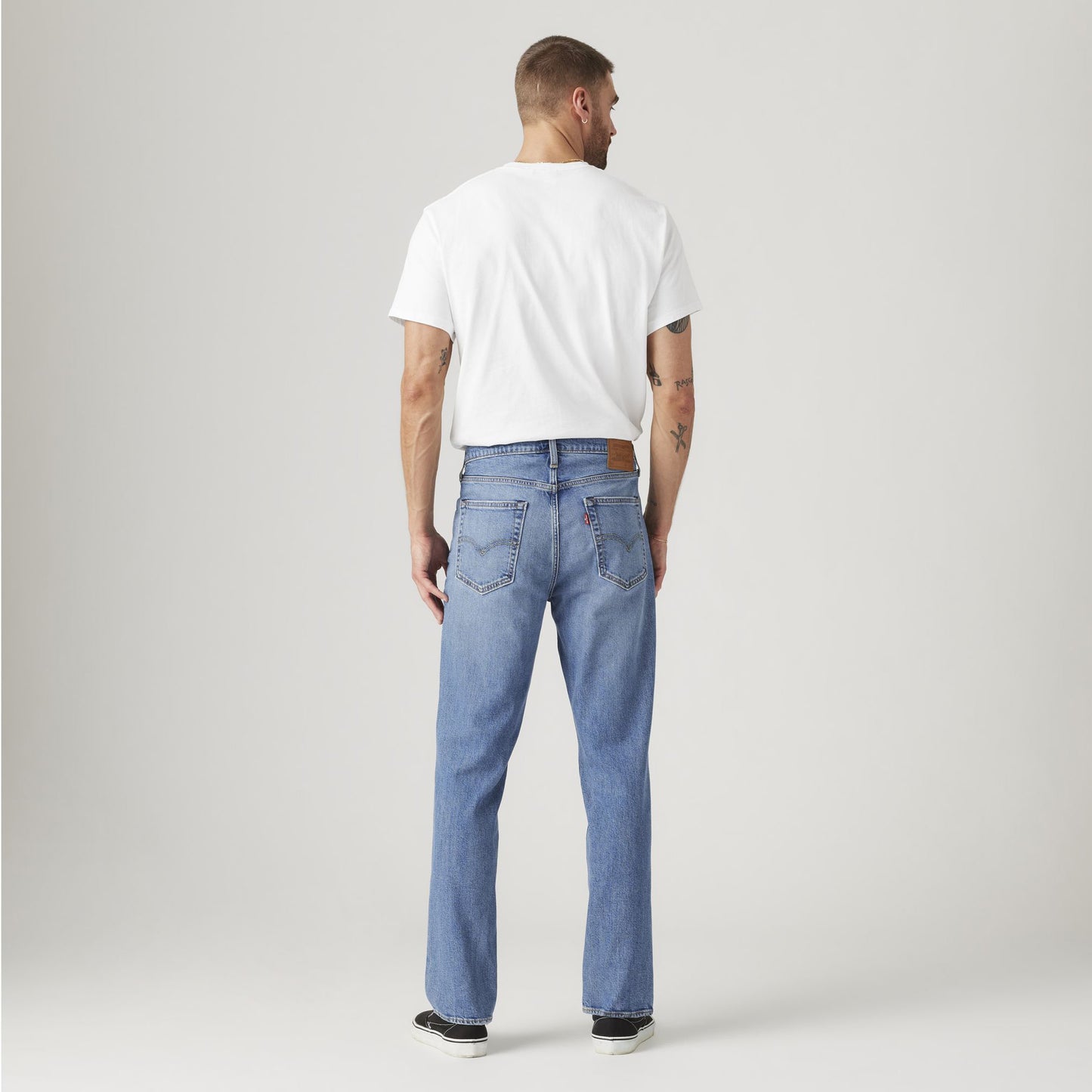A man faces away from the camera, wearing a white T-shirt, Levis 541 Athletic Taper light blue jeans with Stretch Technology, and black sneakers against a plain light background.