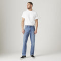 A man stands facing left, wearing a plain white t-shirt, Levi’s 541 Athletic Taper jeans designed with Flex stretch for athletic builds, and black slip-on shoes, against a light gray background.