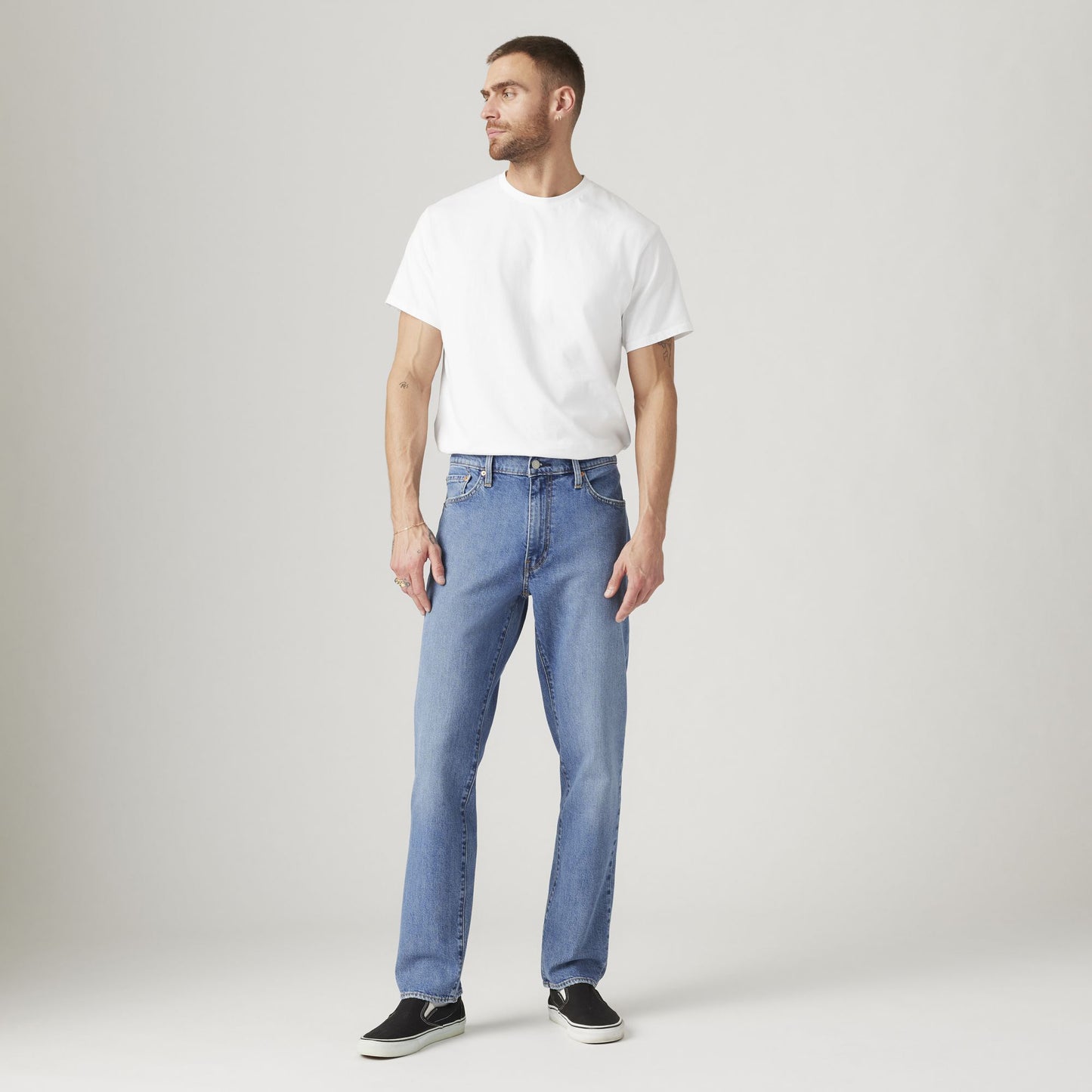 A man stands facing left, wearing a plain white t-shirt, Levi’s 541 Athletic Taper jeans designed with Flex stretch for athletic builds, and black slip-on shoes, against a light gray background.