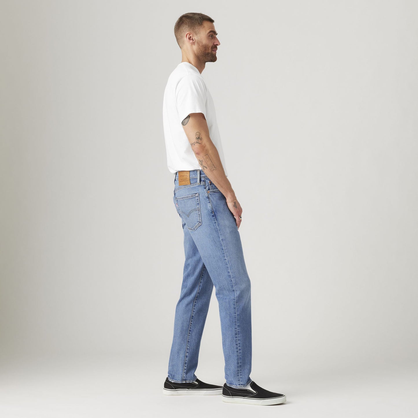 A man stands sideways in a studio wearing a white t-shirt, black slip-on shoes, and light blue Levi's 541 Athletic Taper jeans designed for athletic builds.