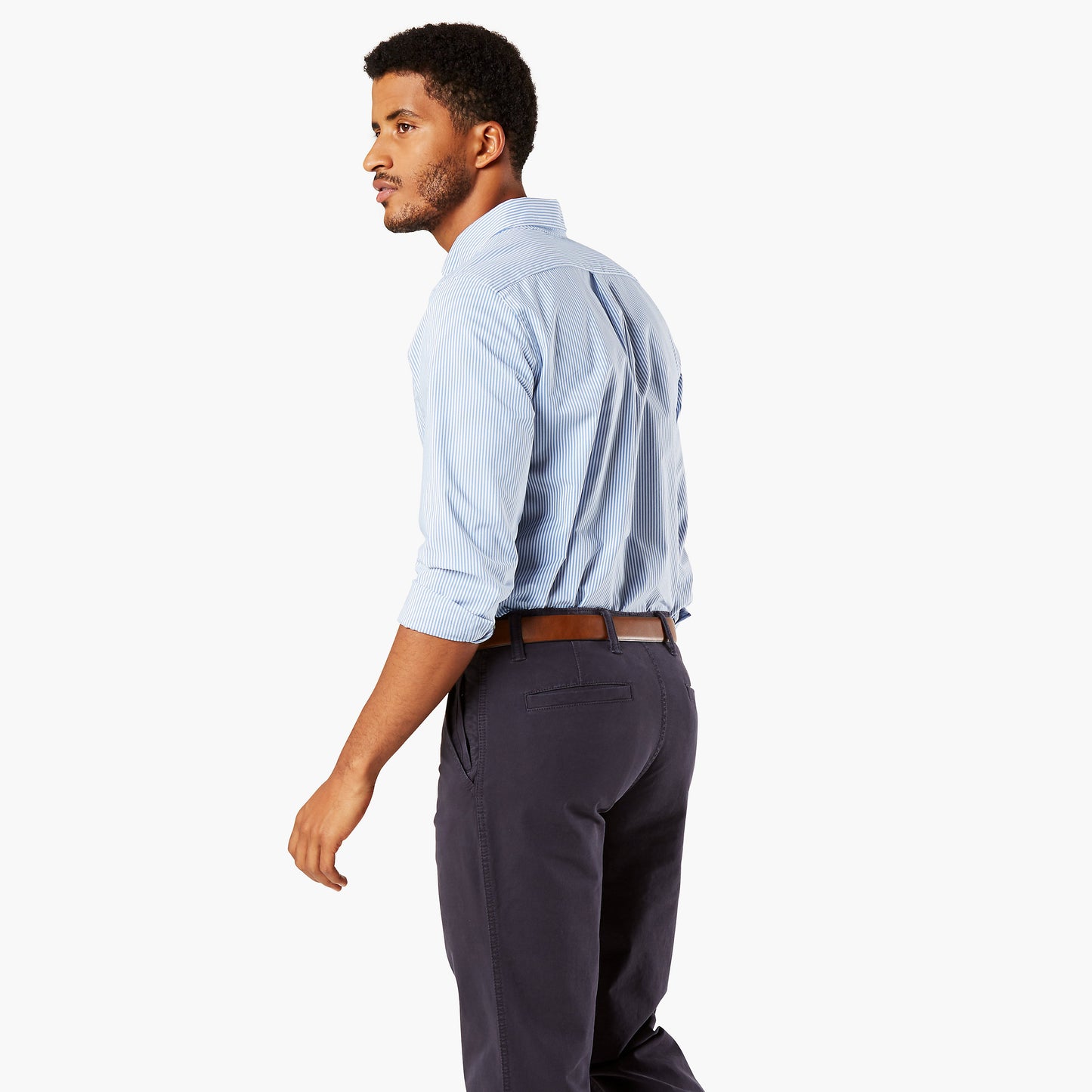 A man in a light blue Dockers Men's Signature Comfort Flex Shirt, LS with a Comfort Flex Collar, dark pants, and a brown belt walks and looks to the side against a plain background.