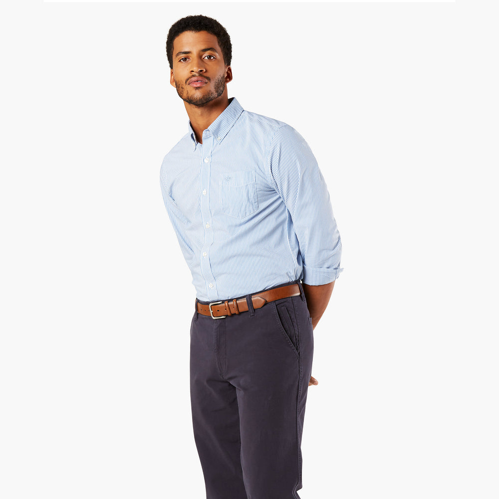 A man wearing a Dockers Men's Signature Comfort Flex Shirt, LS in light blue, paired with dark pants and a brown belt, stands against a plain white background.
