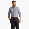 A man wears the Dockers Men's Signature Comfort Flex Shirt, LS in blue check, navy dress pants, and a brown belt, standing with one hand in his pocket against a plain white background.