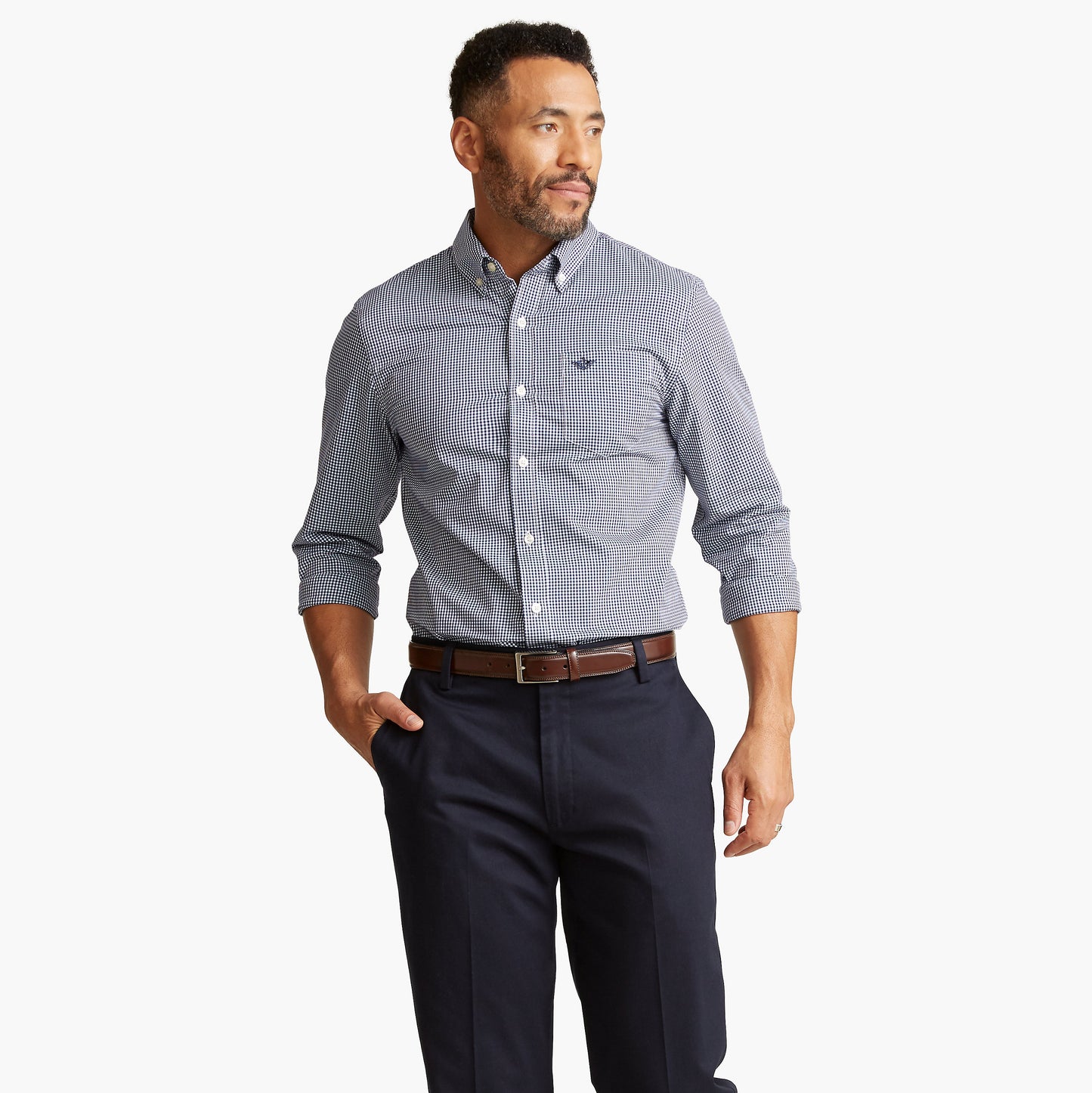 A man wears the Dockers Men's Signature Comfort Flex Shirt, LS in blue check, navy dress pants, and a brown belt, standing with one hand in his pocket against a plain white background.