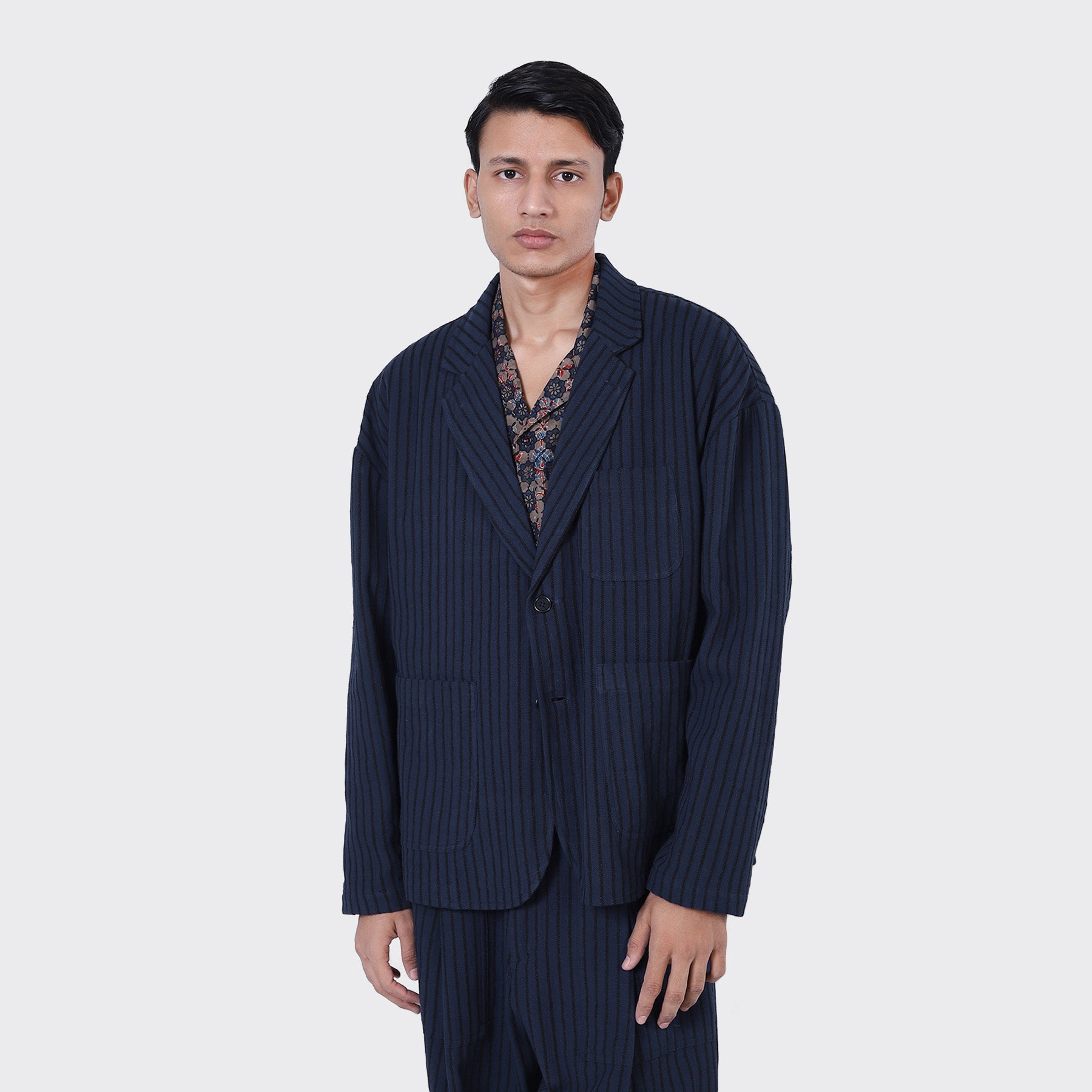 A man stands facing forward against a plain light gray background, wearing Kardo’s Hugh 2.0 Blazer in Blue Black Stripe over a patterned shirt.