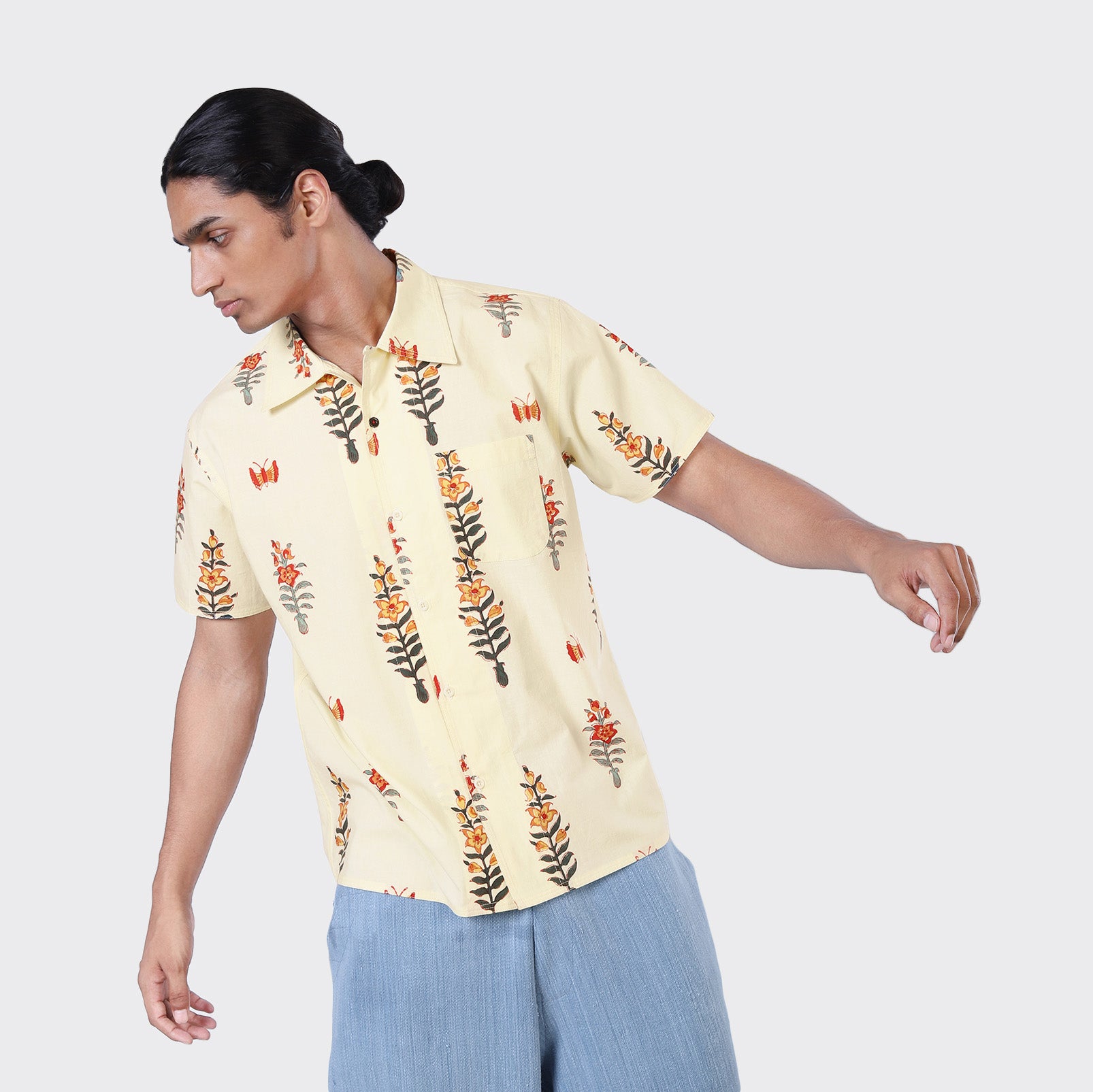 Wearing the Kardo Chintan SS Shirt, Block Print 138—a light yellow, floral-print short-sleeve shirt—and light blue pants, a person stands against a plain background, looking to the side with one arm outstretched.