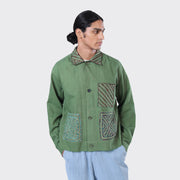 A person in the Kardo Bodhi Jacket, Matcha Rabari Applique—featuring green fabric with patterned pockets and collar—and light blue pants, stands against a plain light background.