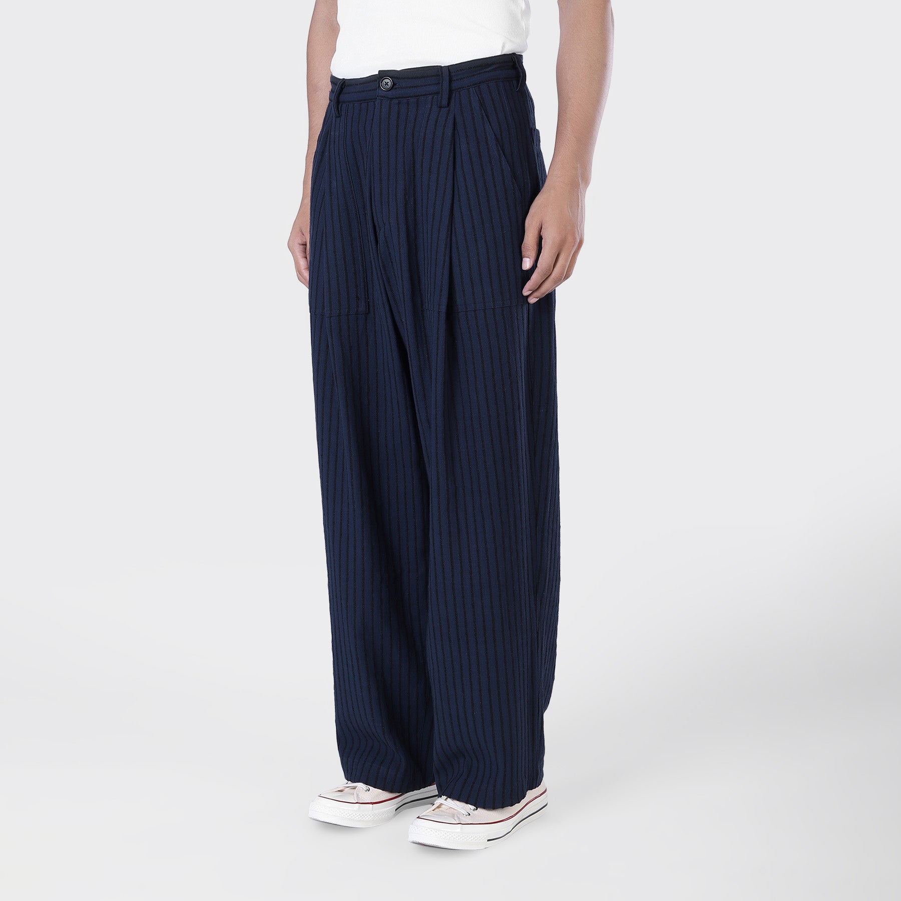 A person stands against a plain light background, wearing a white shirt, white sneakers, and Kardo Yuki Pleated Trousers in Blue Black Stripe.