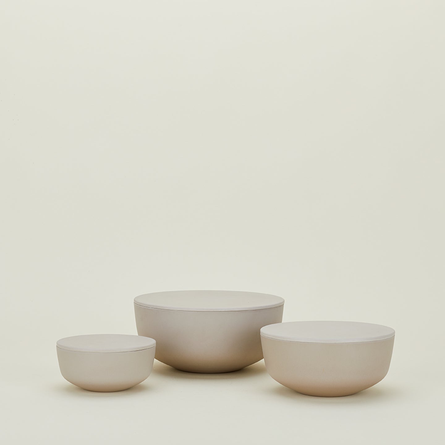 The "Essential Lidded Bowls" set of three beige ceramic bowls by Hawkins New York, designed for stackable storage, is arranged on a plain, light background.