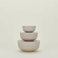 The Essential Lidded Bowls set by Hawkins New York features three stackable, minimalist beige ceramic bowls of varying sizes resting against a plain light background. Their elegant design makes them ideal for any modern kitchen or dining space.