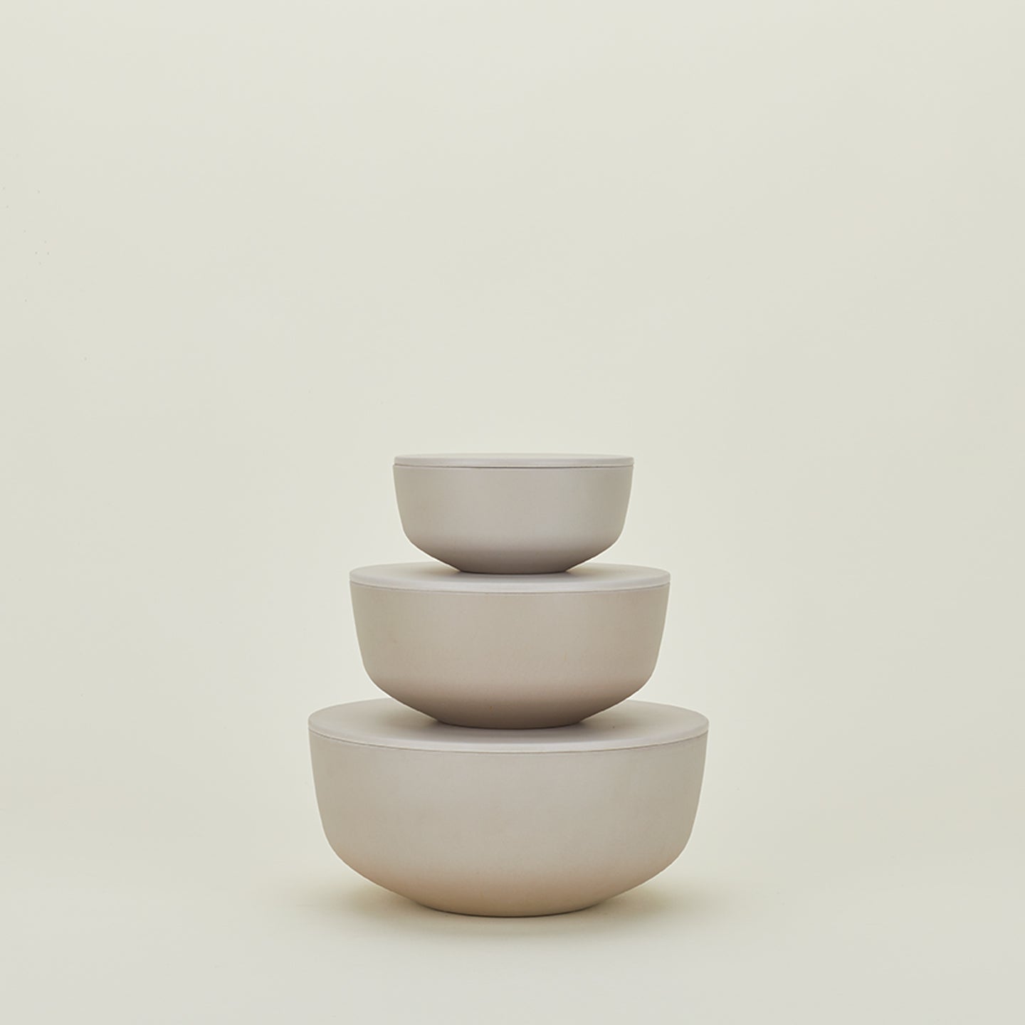 The Essential Lidded Bowls set by Hawkins New York features three stackable, minimalist beige ceramic bowls of varying sizes resting against a plain light background. Their elegant design makes them ideal for any modern kitchen or dining space.