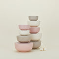 A set of pastel Essential Lidded Bowls by Hawkins New York, crafted from recycled bamboo, is neatly stacked with varying sizes. Beside them, two garlic bulbs rest on a light background for a rustic touch.