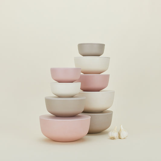A set of pastel Essential Lidded Bowls by Hawkins New York, crafted from recycled bamboo, is neatly stacked with varying sizes. Beside them, two garlic bulbs rest on a light background for a rustic touch.