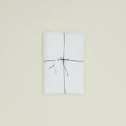 A neatly folded Hawkins New York Simple Linen Bedding - Queen Flat Sheet in white, tied with black string and a white tag, rests on a plain light background. Woven in Portugal for timeless quality.