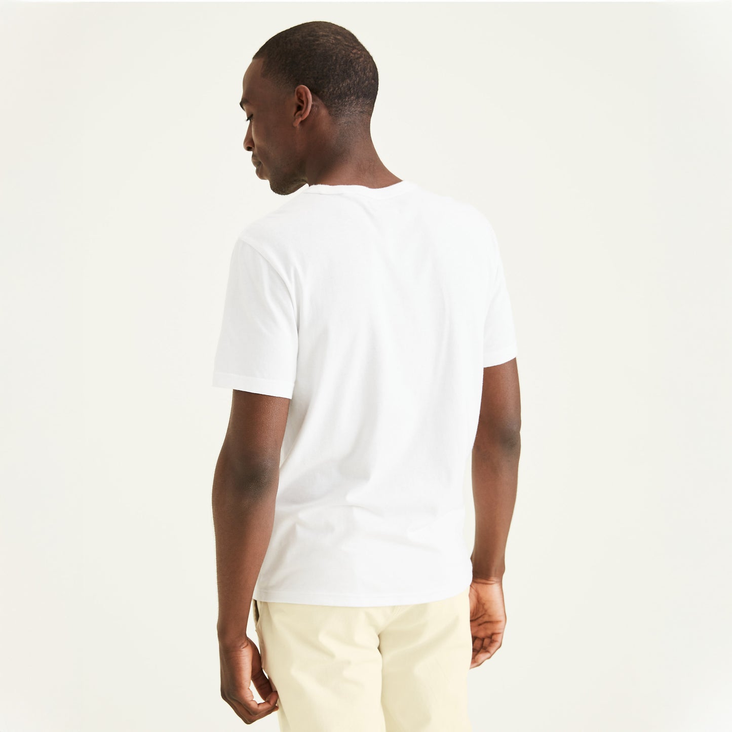 A man stands with his back to the camera, wearing a Dockers Men's Crewneck Tee in slim fit plain white and light beige pants, set against a plain light background.