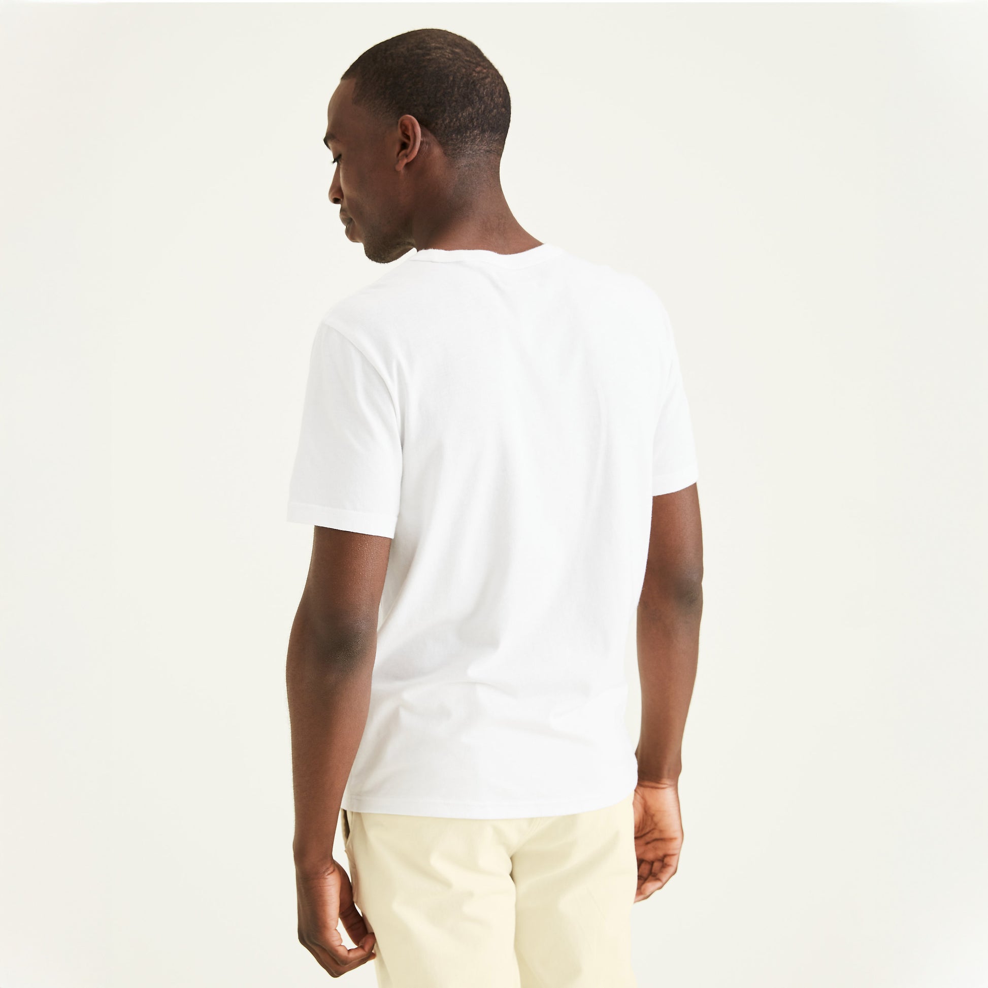 A man stands with his back to the camera, wearing a Dockers Men's Crewneck Tee in slim fit plain white and light beige pants, set against a plain light background.