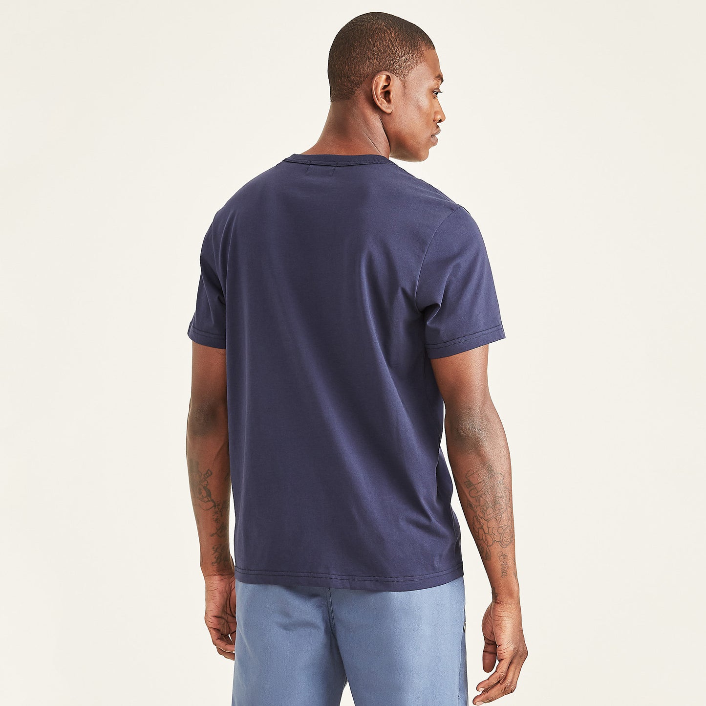 A man stands facing away against a plain light background, wearing the Dockers Men's Crewneck Tee in navy blue slim fit and light blue pants, highlighting the back view of his outfit.