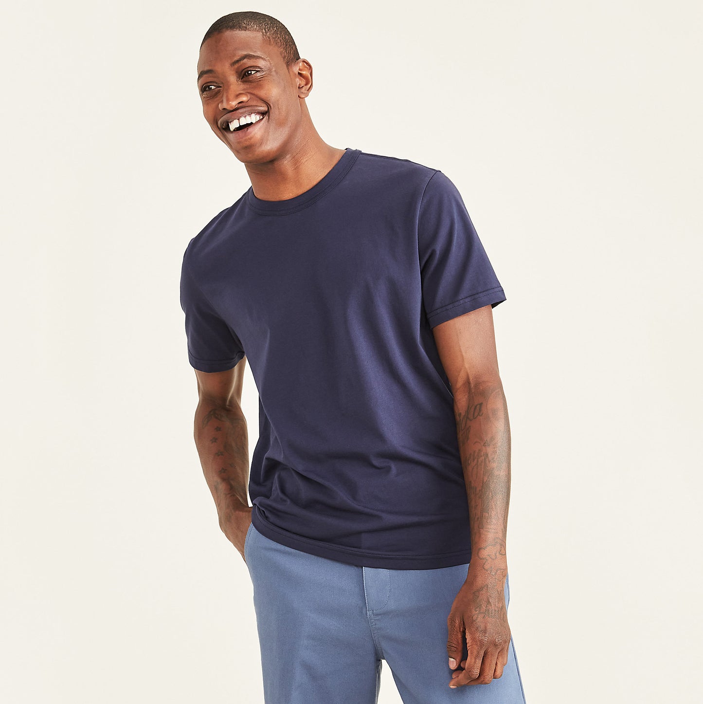 A man stands smiling against a plain light background, wearing a Dockers Men's Crewneck Tee in slim fit navy blue paired with light blue pants.