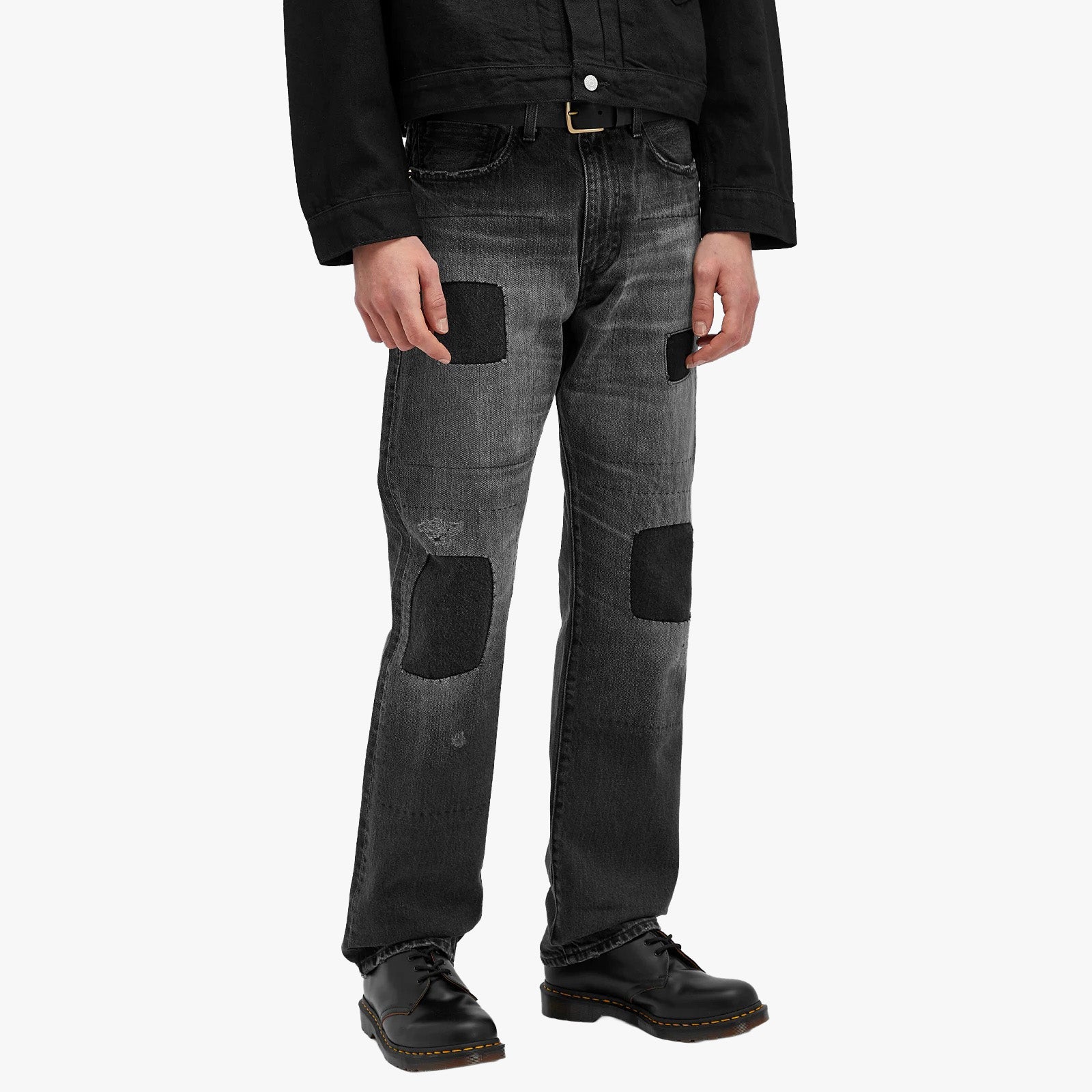 A person wearing Levis 505 MIJ Texas Tea black patched gray jeans, a black jacket, and black shoes stands against a plain light background.