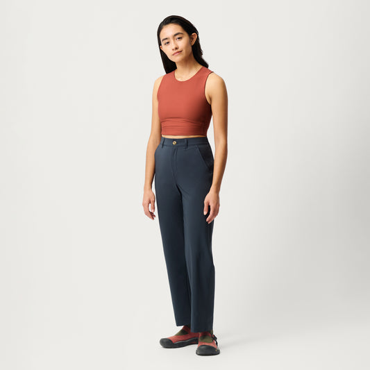 A woman stands against a plain background, wearing a sleeveless rust-colored crop top, comfortable Hikerkind Trail Trousers 02 in dark blue, and dark shoes with red socks.
