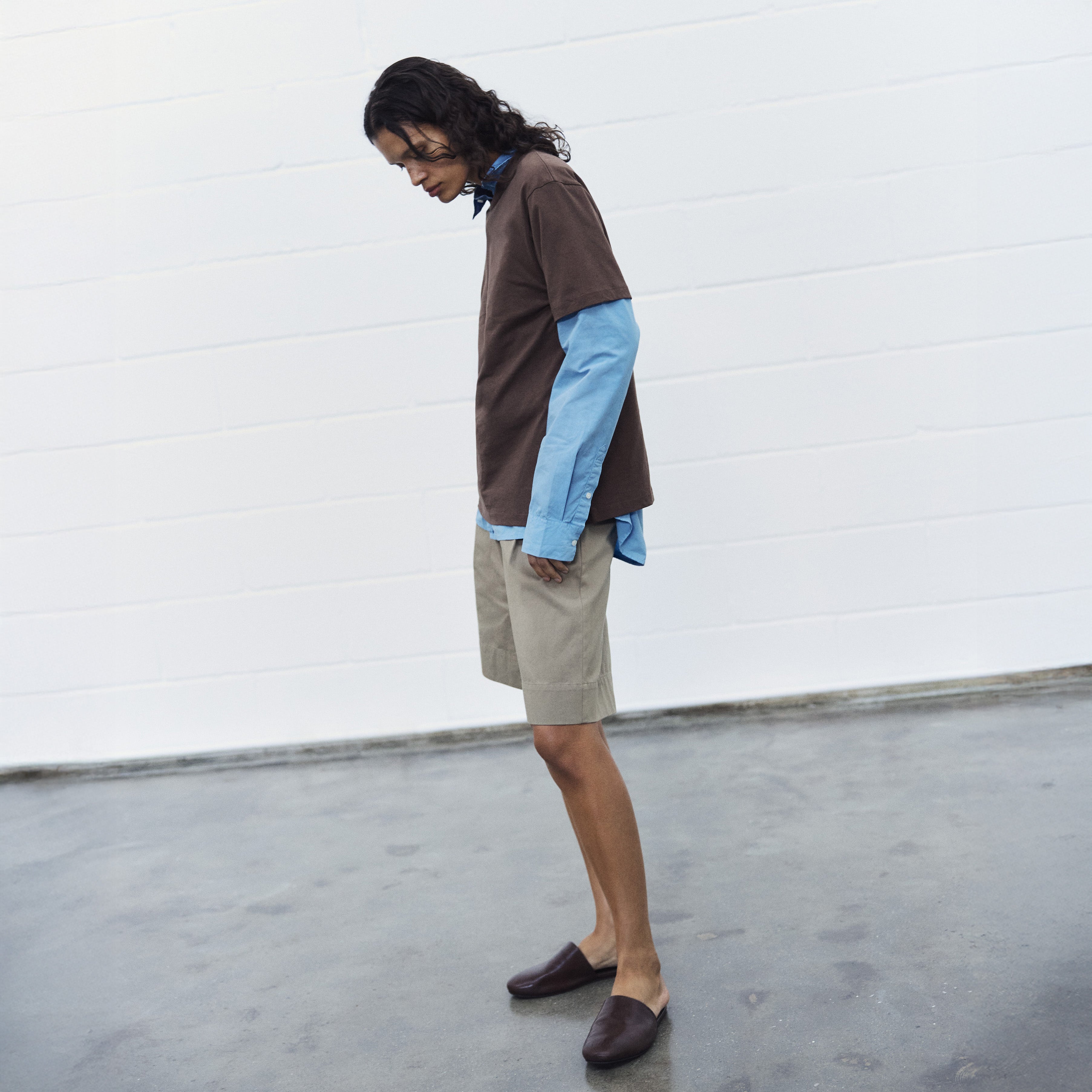 Indoors, a person wears the Aiayu Classic Circular Tee II over a blue long sleeve shirt, paired with beige shorts and brown slip-on shoes, looking down.