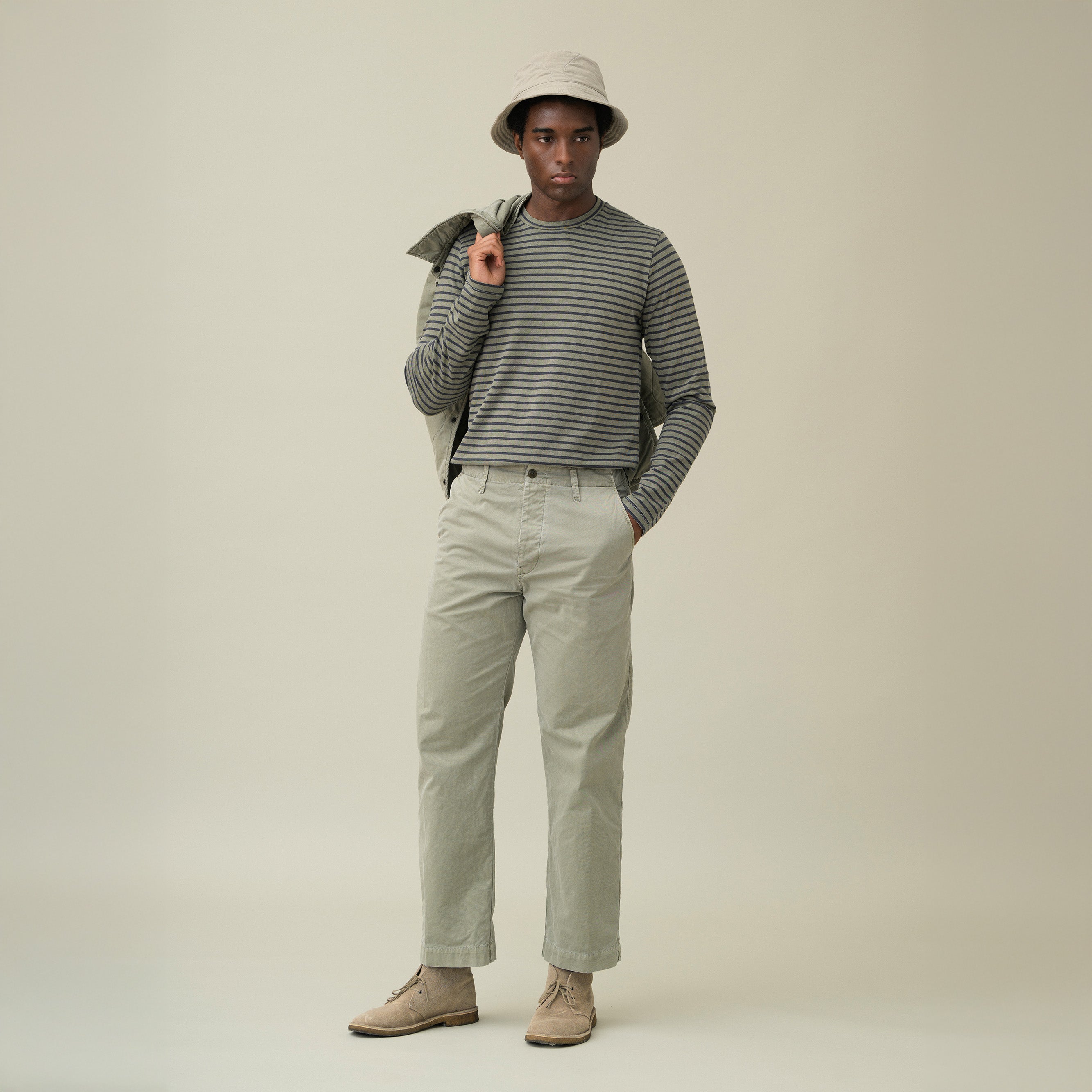 A man stands in a neutral studio wearing a striped long-sleeve shirt, beige boots, a light bucket hat, and Save Khaki’s Weekend Chino in khaki, holding a jacket over his shoulder.