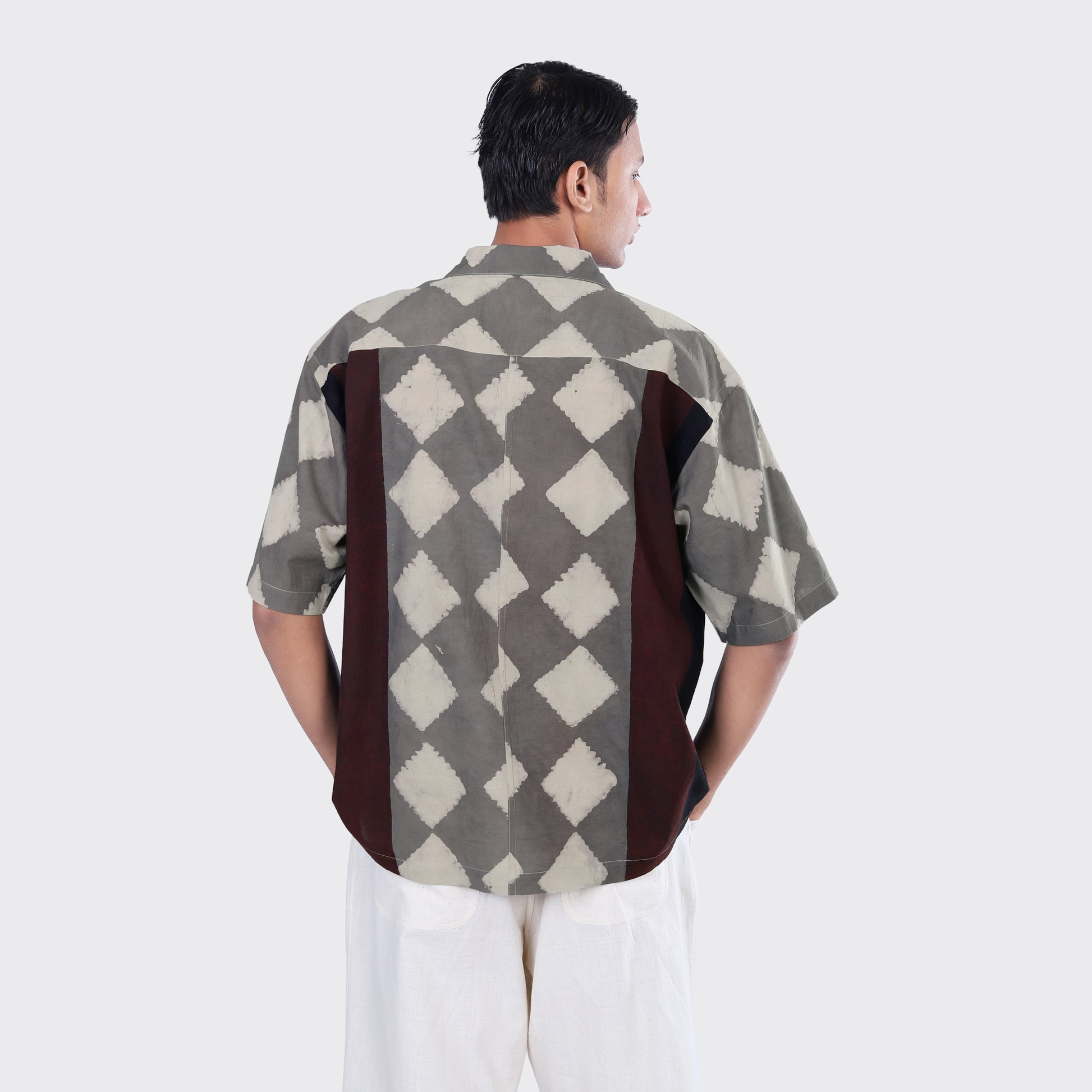 A man facing away wears the Kardo Rohit Shirt, Block Print 139—a short-sleeve shirt with a gray and white diamond pattern and dark side panels—paired with light-colored pants.