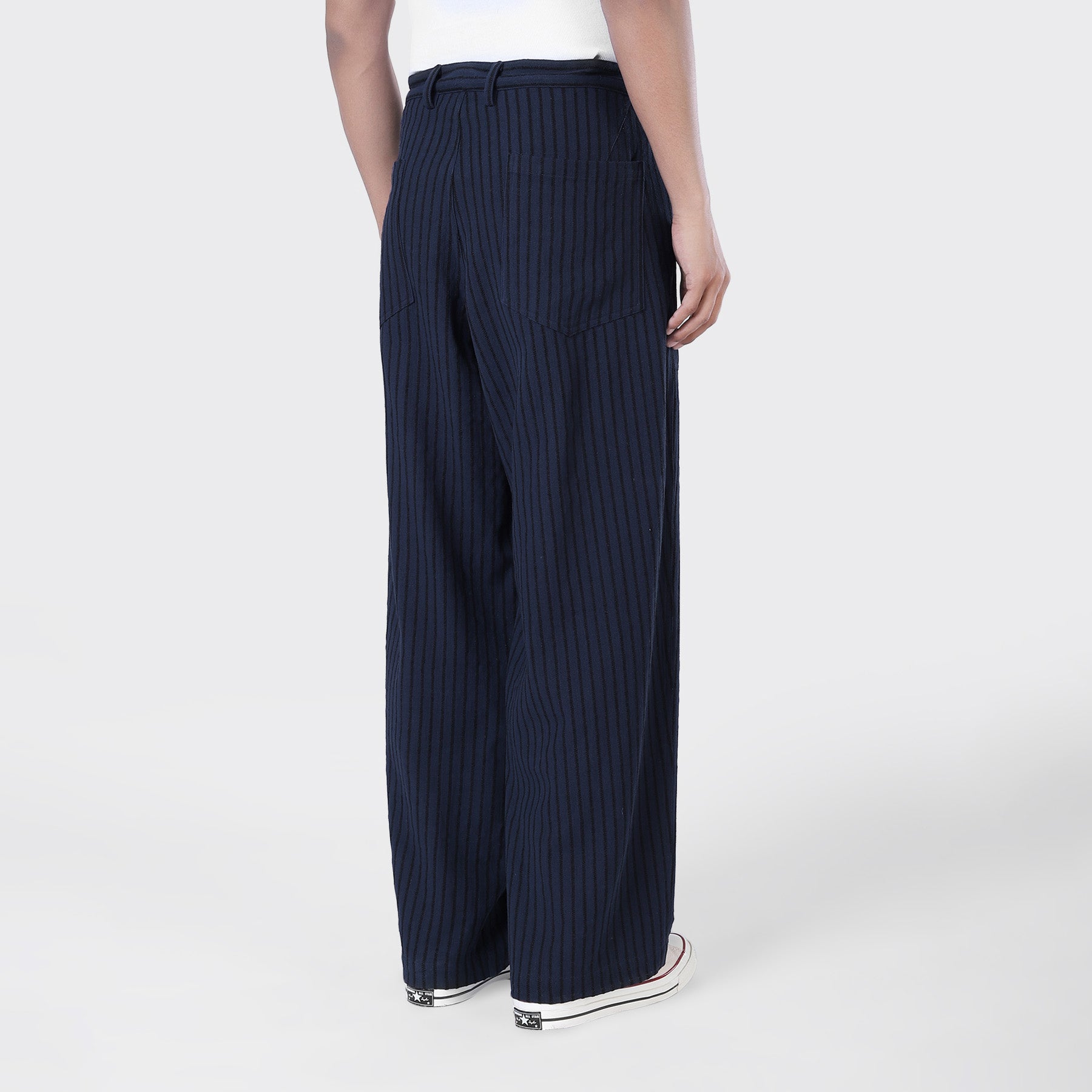 Person wearing Kardo’s Yuki Pleated Trousers in Blue Black Stripe, paired with white sneakers, shown from the back against a plain light background.