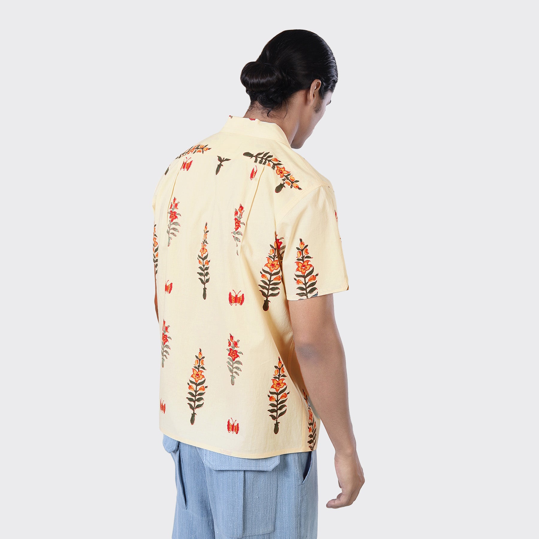A person with long dark hair in a low bun faces away, wearing the Kardo Chintan SS Shirt in Block Print 138—a light yellow short-sleeve shirt with a red and brown floral pattern—paired with light blue pants.