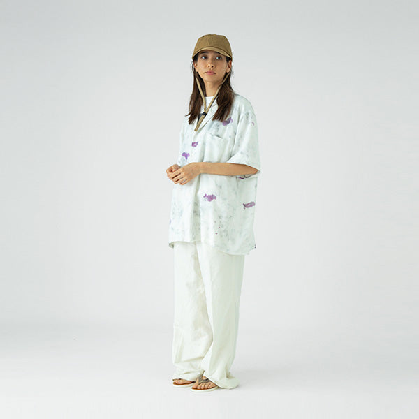 A woman poses against a plain background wearing the Sublime Roe Charidoor Cap, paired with a loose white patterned shirt, wide white pants, and sandals.