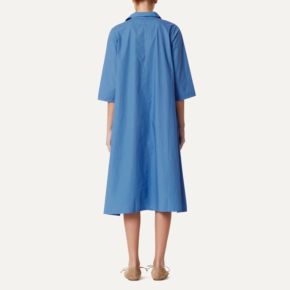 Individual wearing the Clara Abito Pepe Dress by Labo Art, a knee-length blue poplin design with elbow-length sleeves and a loose fit for a casual yet elegant appearance, paired with light brown shoes, facing away.