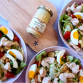 Three bowls of salad with eggs, cherry tomatoes, and chicken surrounding a jar of Edmund Fallot Dijon Mustards from Gourmet Food Solutions, Inc. on a wooden cutting board with a star cutout.