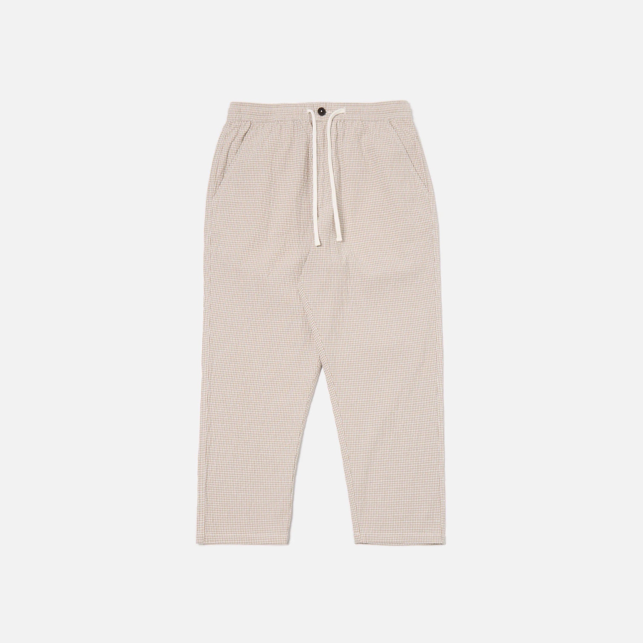 The Ospina Check Cotton High Water Trouser in Ecru/Sand by Universal Works features a button closure, elastic waistband, side pockets, and straight leg fit, shown laid flat on a white background.