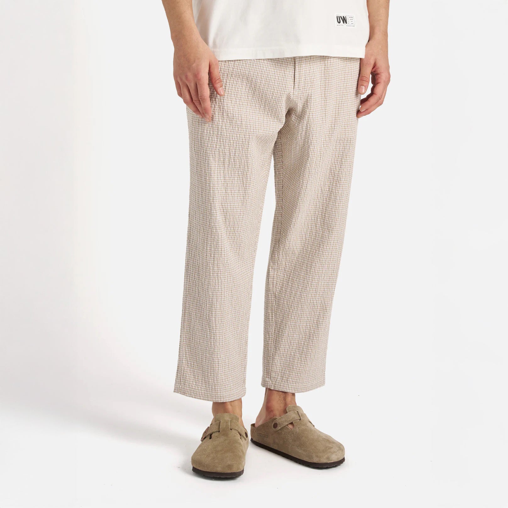 A person wears Universal Works' Ospina Check Cotton High Water Trouser in Ecru/Sand with a white shirt and taupe slip-on shoes, standing against a plain white background.